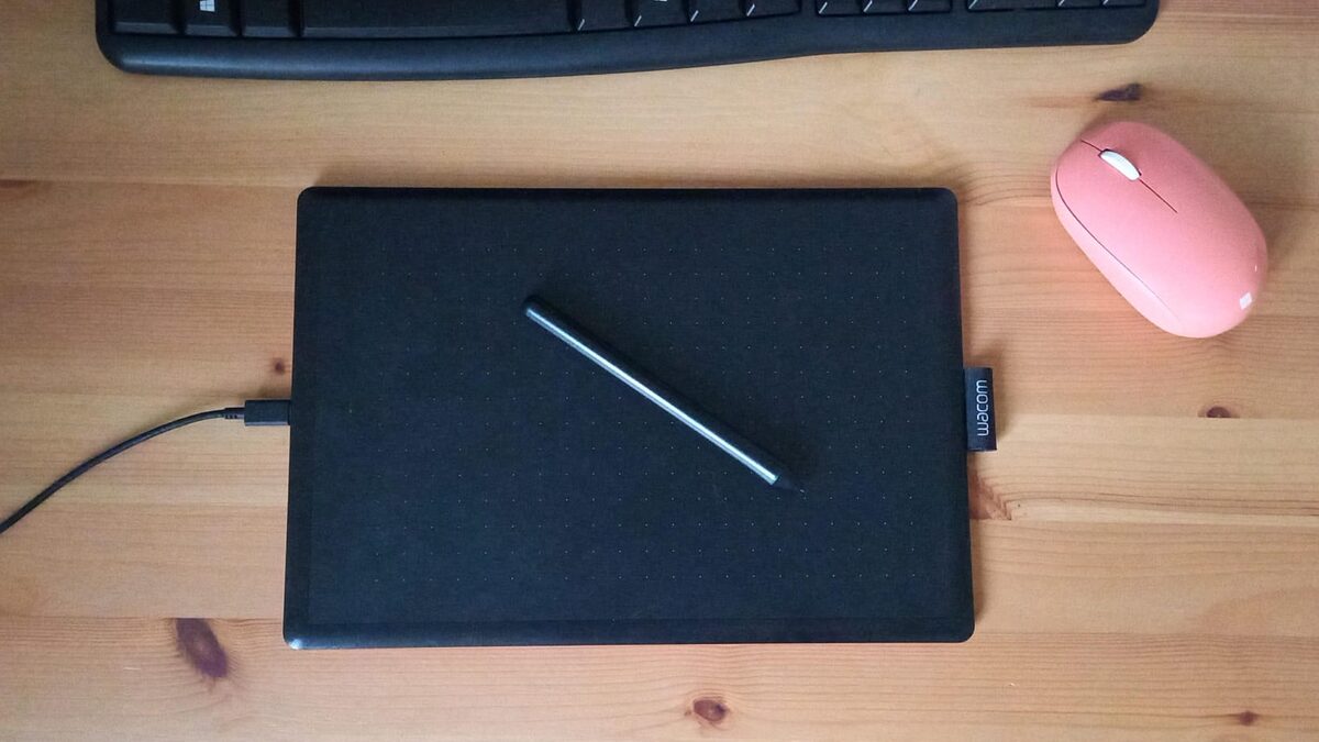 A graphics tablet and stylus beside a keyboard and pink mouse on a wooden desk.