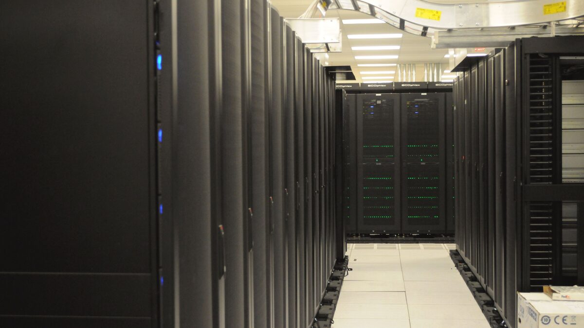 An aisle of black server racks with overhead cable trays inside a working data centre.